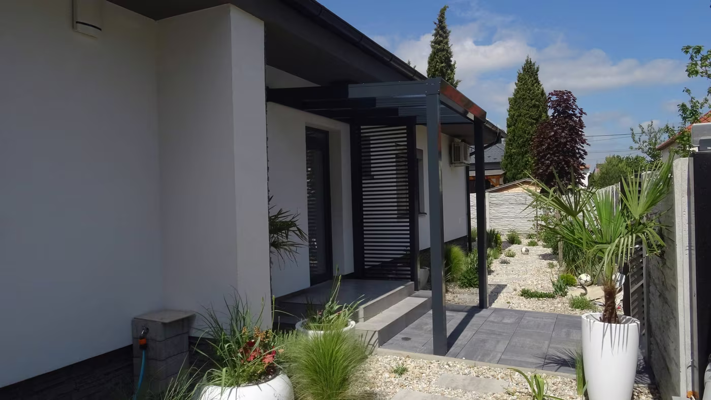 Antracitová hliníková pergola Economic Class pozdĺž bieleho domu s kvetináčmi