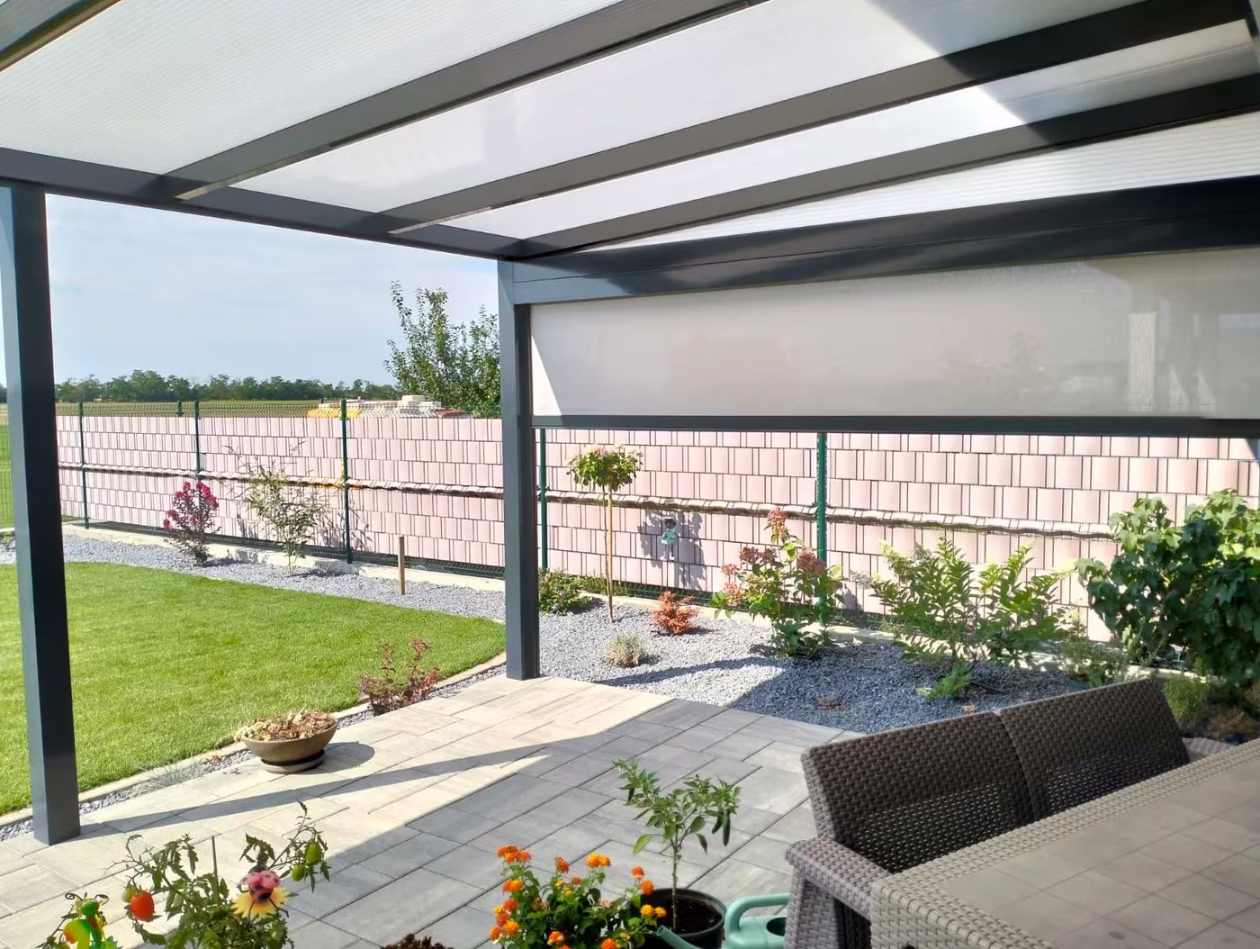 Antracitová hliníková pergola Economic Class pozdĺž plota s ratanovým sedením a kvetmi