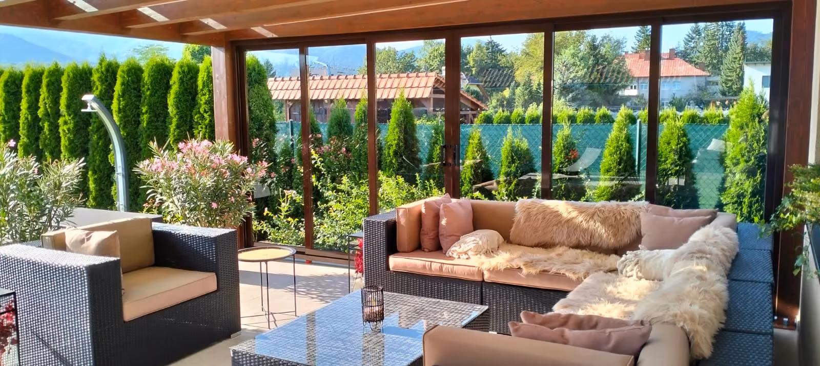 Zariadená drevená pergola Milano s veľkou sedačkou pri dome s červenou strechou