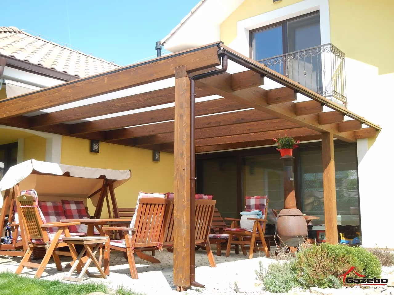 Drevená pergola Fénix nad terasou so záhradným nábytkom a hojdačkou pri žltom dome