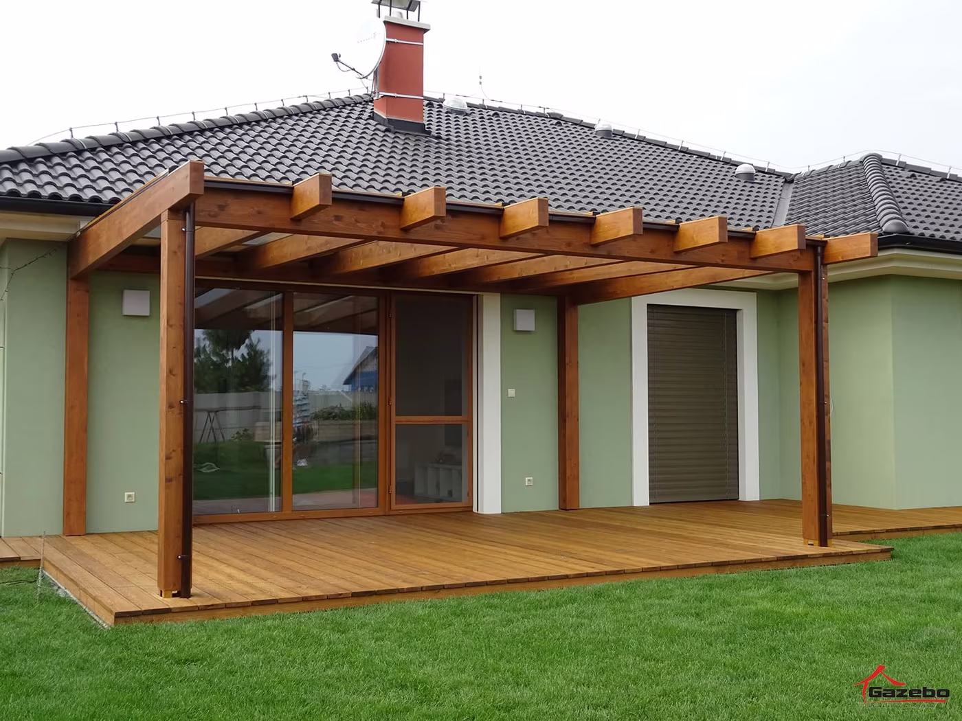 Drevená pergola Fénix pri dome s presklenými dverami nad drevenou terasou s trávnikom